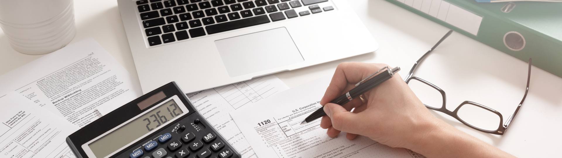 Hand filling out tax papers on a desk with a calculator and laptop